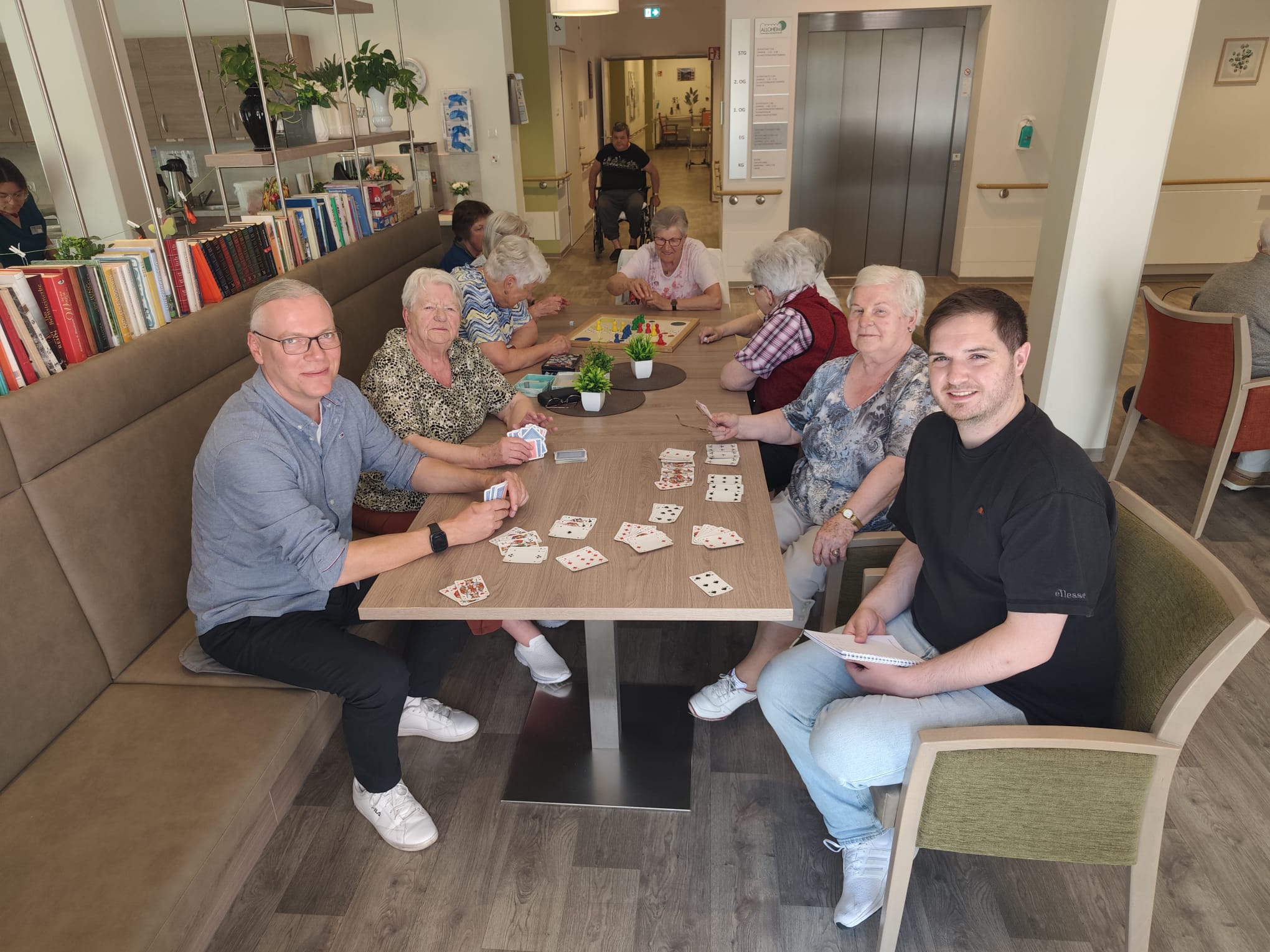 Eine Gruppe Seniorinnen und junge Männer sitzt an einem Tisch und spielt Karten.